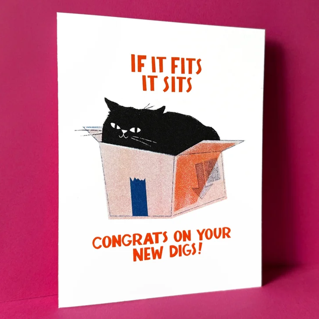 A white greeting card featuring a black cat sitting in a cardboard box with orange text reading 'IF IT FITS IT SITS' and 'CONGRATS ON YOUR NEW DIGS!', photographed standing against a magenta-pink background.