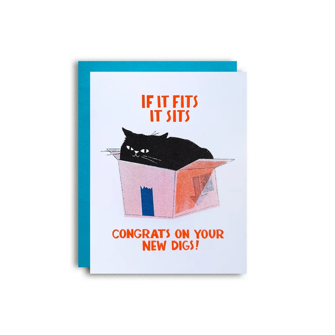 A novelty greeting card with a white front featuring an illustration of a black cat sitting in a pink box, with orange text reading 'IF IT FITS IT SITS' and 'CONGRATS ON YOUR NEW DIGS!'. A bright blue envelope is partially visible behind the card on a white background.