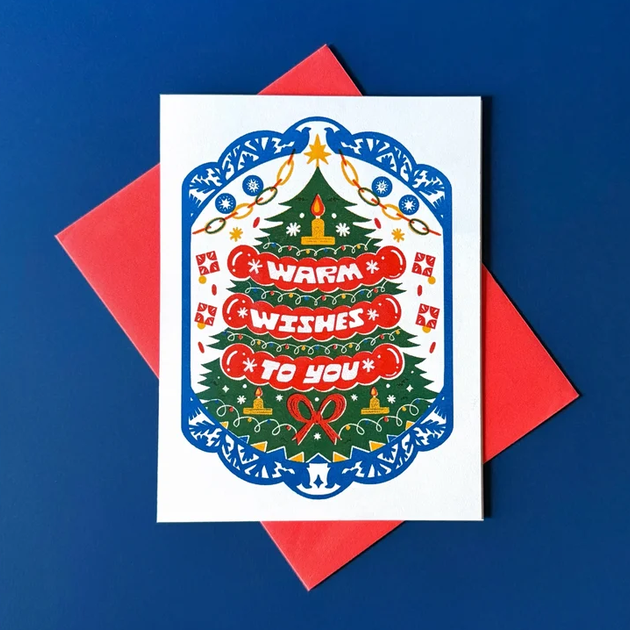 A square white Christmas greeting card with a colorful illustrated tree and the words “WARM WISHES TO YOU” in red, placed on top of a red envelope against a blue background.
