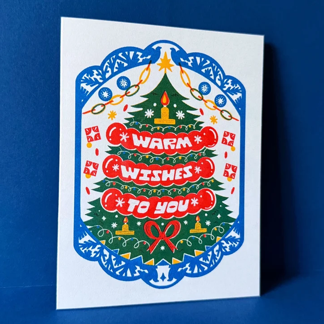 A vertical greeting card with a colorful Christmas tree illustration and the words 'WARM WISHES TO YOU' in red, shown standing against a dark blue background.