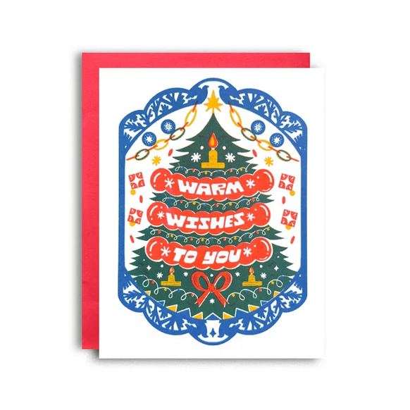 A Christmas greeting card with a colorful folk-art style illustration of a decorated tree and the words 'WARM WISHEST TO YOU' in red banners, shown with a red envelope behind it on a white background.
