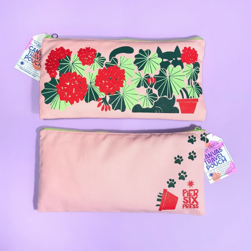 Two pastel pink canvas travel pouches with green zipper trim are displayed on a lavender background. The top pouch features a colorful graphic print of a black cat among red flowers and green leaves, with a hanging tag reading 'CANVAS TRAVEL POUCH' and 'CANVAS PRESS'.