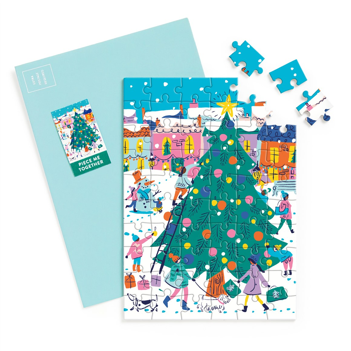 A Christmas-themed jigsaw puzzle with a light blue envelope, shown with several loose puzzle pieces on a white background. The puzzle features a festive winter village scene with a large decorated Christmas tree in the foreground, colorful houses, snow, and people walking and playing.
