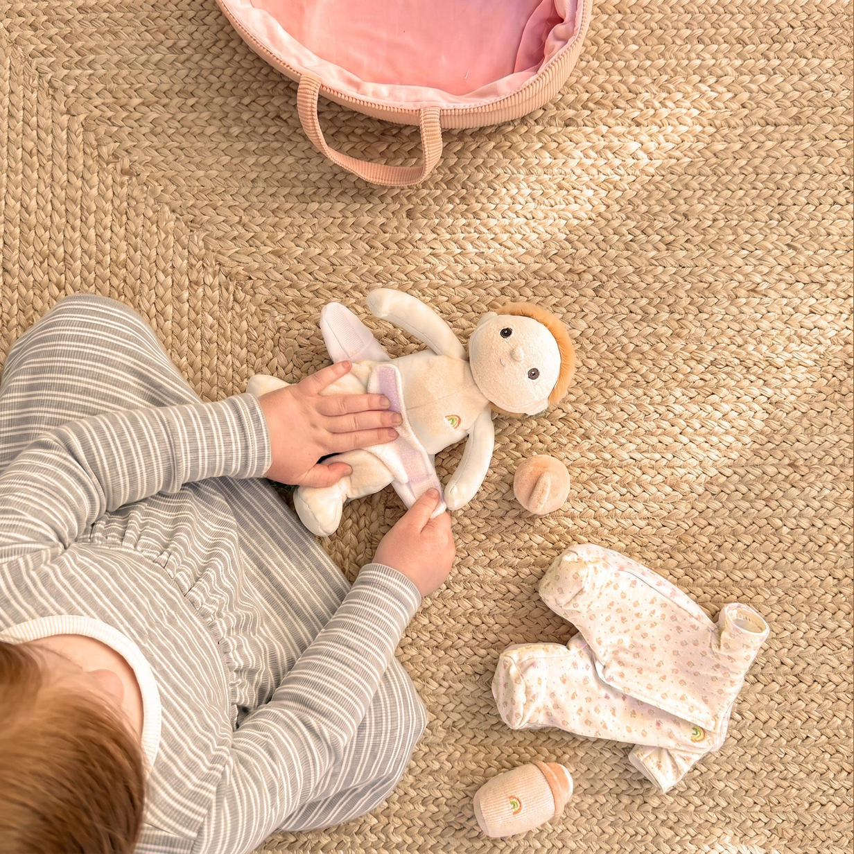 A baby sits on a woven straw rug interacting with a plush monkey toy and a soft clothing set. The baby wears a gray and white striped long-sleeve top.