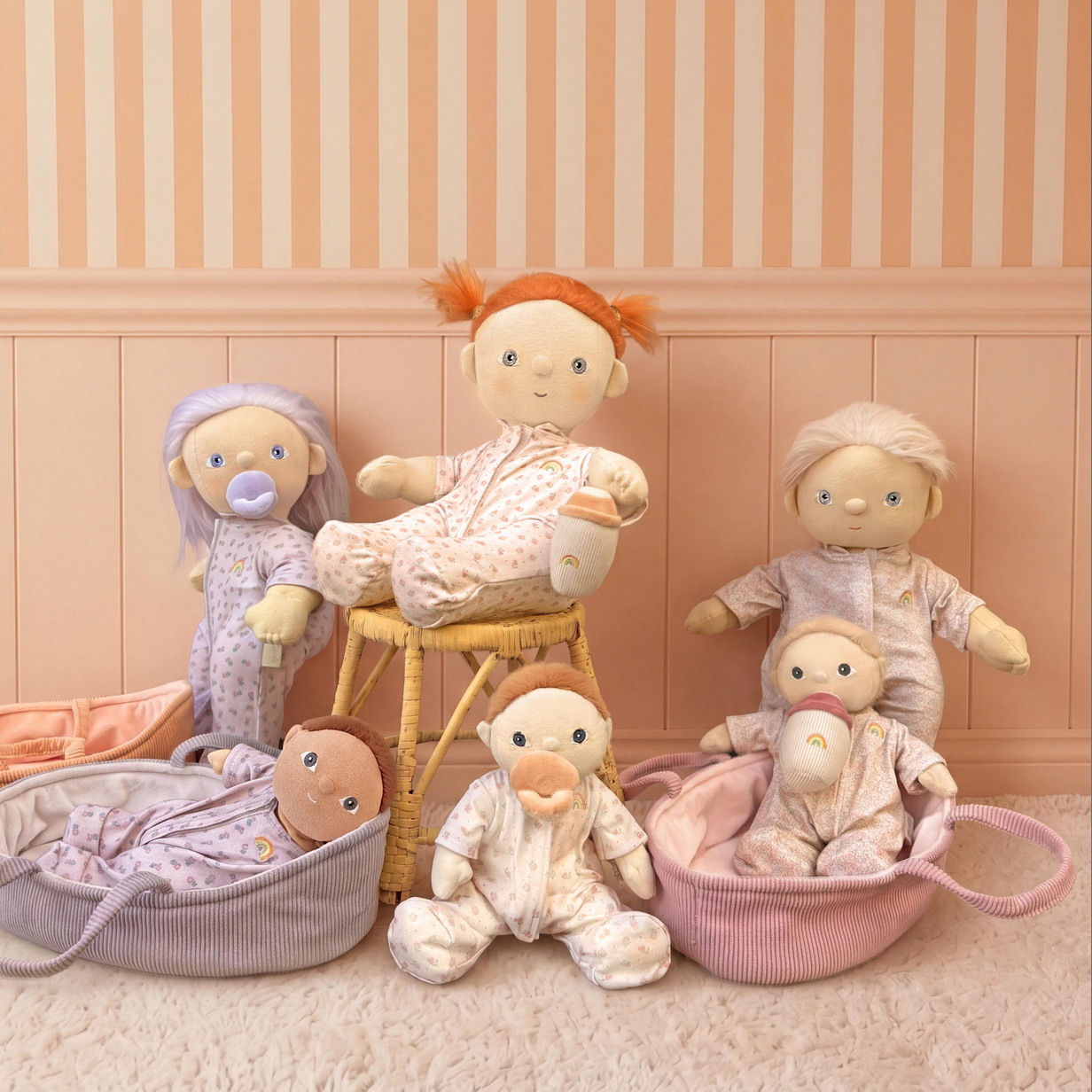 A collection of soft plush baby dolls in coordinated outfits, arranged on a stool and in baskets against a peach-and-cream striped wall.