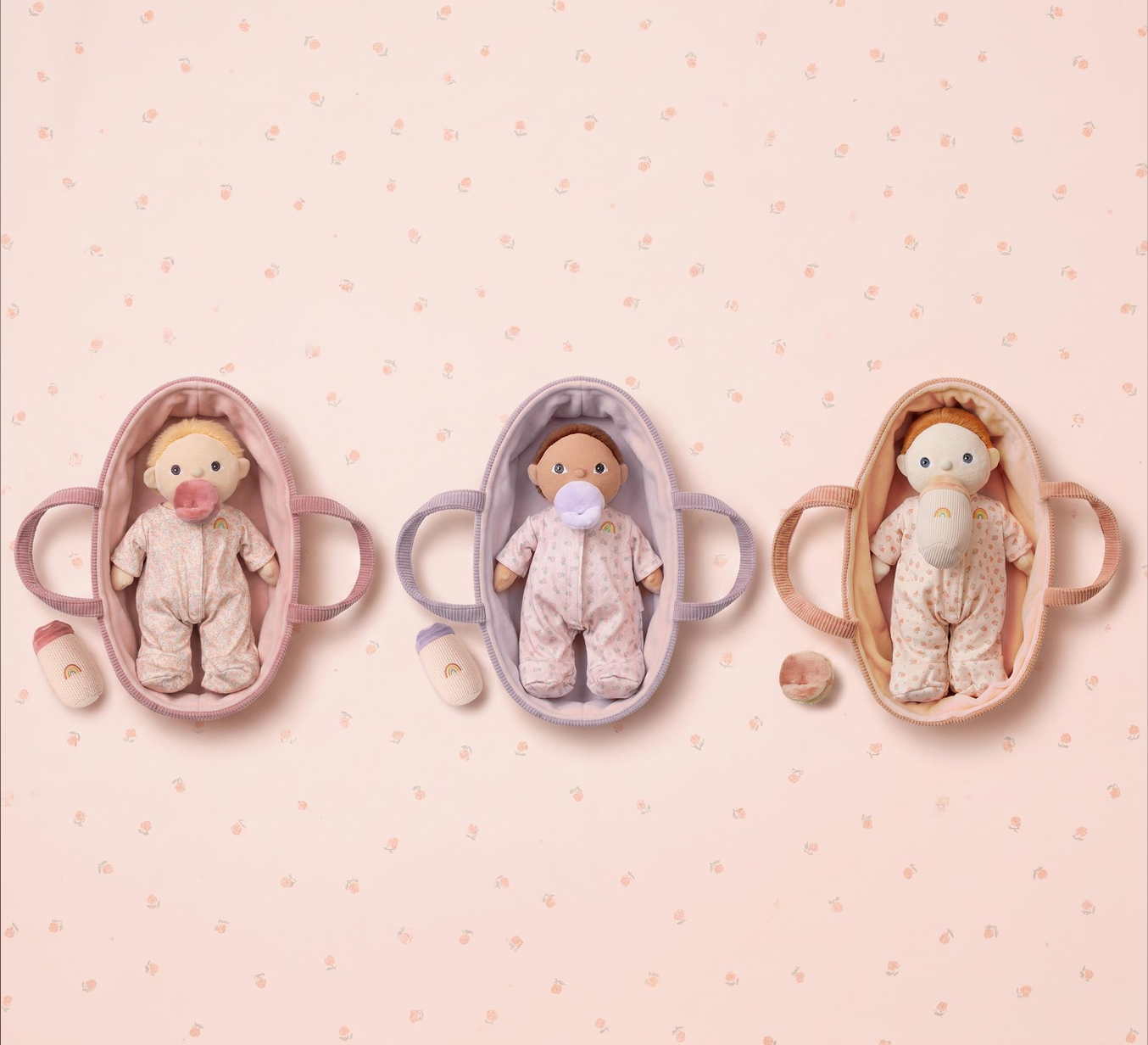 Three baby doll play cribs with handles are arranged on a light pink patterned background. Each crib contains a baby doll wearing a matching pink and white patterned onesie, with a pacifier and small accessories beside them.