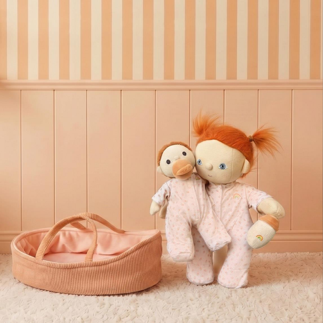 A soft toy doll with orange hair and a smaller doll with brown hair, both wearing pale pink patterned pajamas, standing beside a peach corduroy bassinet on a cream rug against a peach paneled wall.