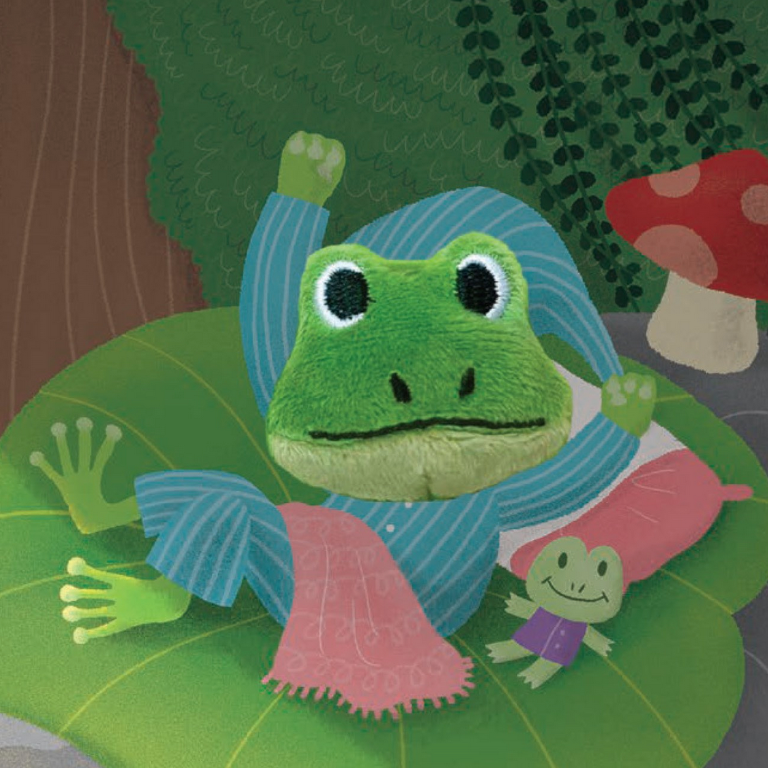 A plush frog toy wearing blue striped pajamas and a pink blanket, lying on a green leaf with a small matching plush frog beside it, set against a whimsical forest background with a red mushroom.