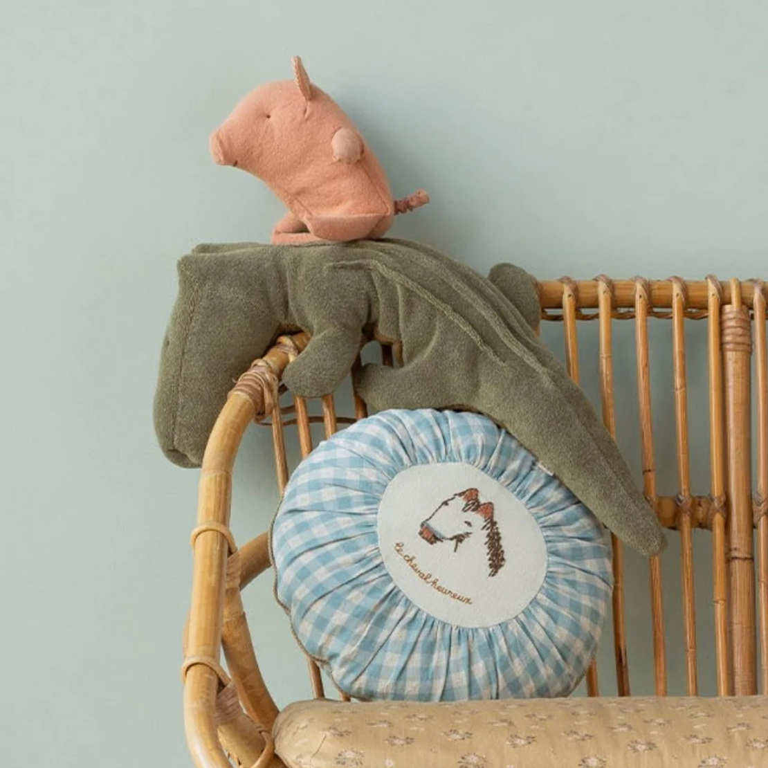 A styled nursery scene featuring a plush pink pig toy, an olive green alligator plush, a round blue-and-white gingham cushion with a white embroidered patch, and a rattan baby chair against a pale blue wall.