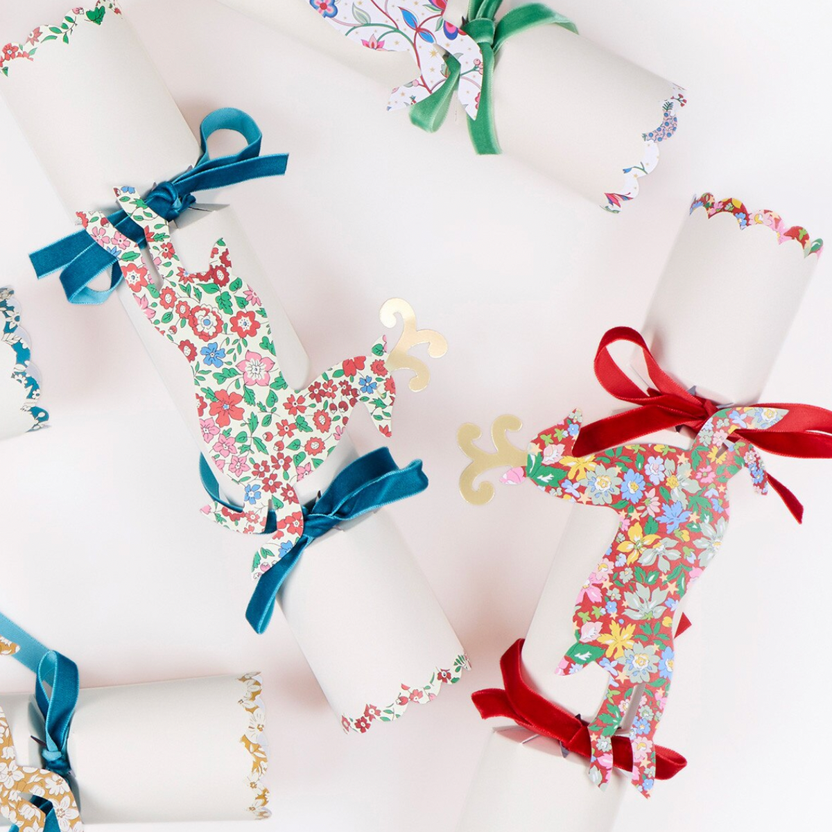 Four white paper Christmas crackers decorated with colorful floral patterns and tied with satin ribbons in blue, green, red, and gold, arranged on a clean white background.