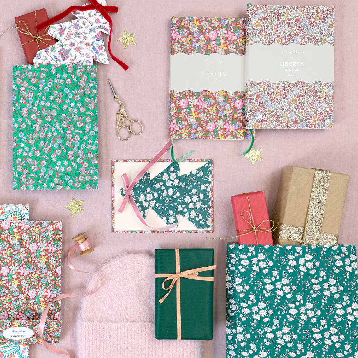 A styled flat-lay of gift wrapping paper and wrapping supplies arranged on a pink surface, including floral patterned sheets, a green floral sheet, kraft paper, ribbon, scissors, and wrapped presents.