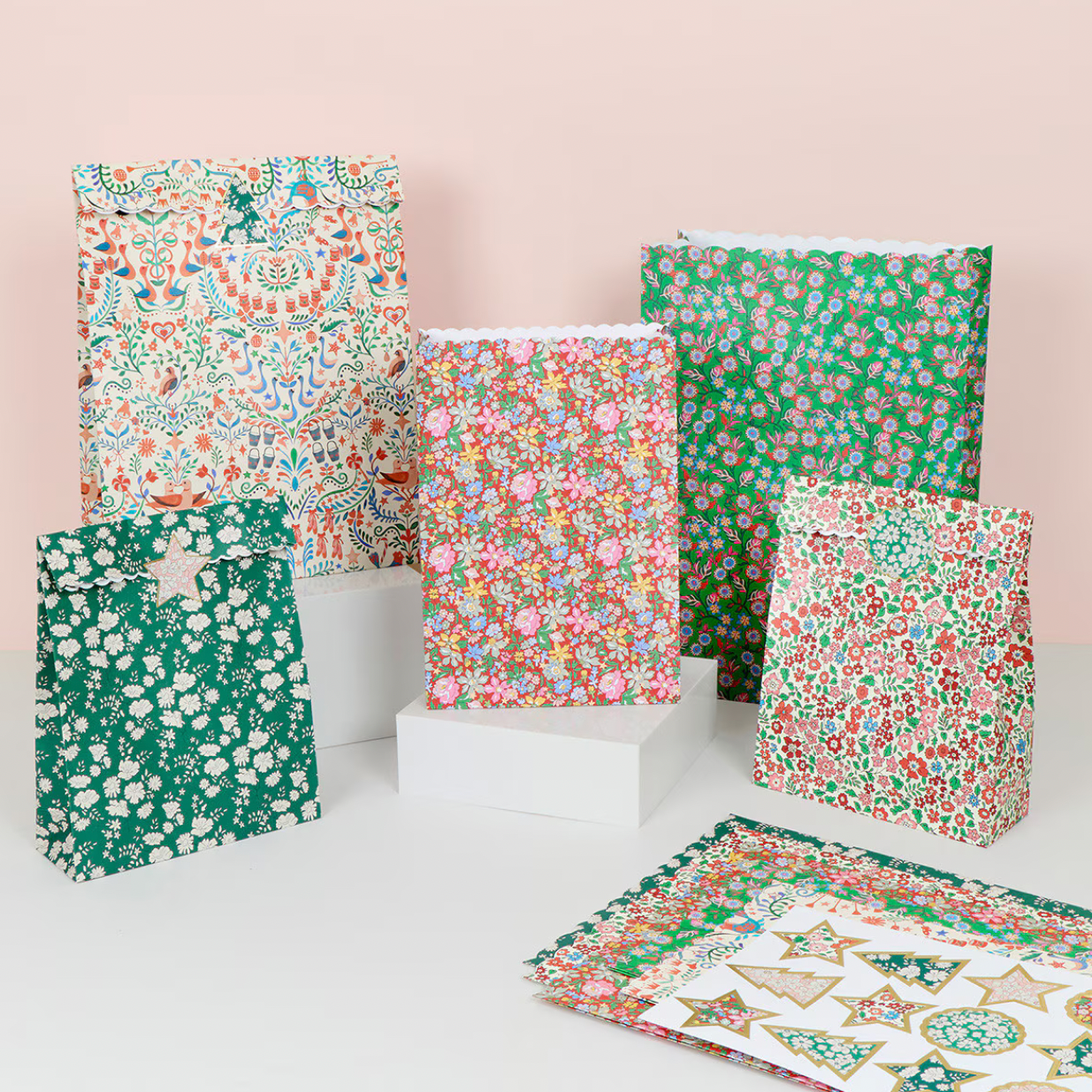 A set of paper gift bags and matching flat paper pieces displayed on white blocks against a pale pink studio background. The bags feature various colorful floral and ornamental patterns in green, pink, red, blue, and white, with one bag showing a white floral print on a green base.