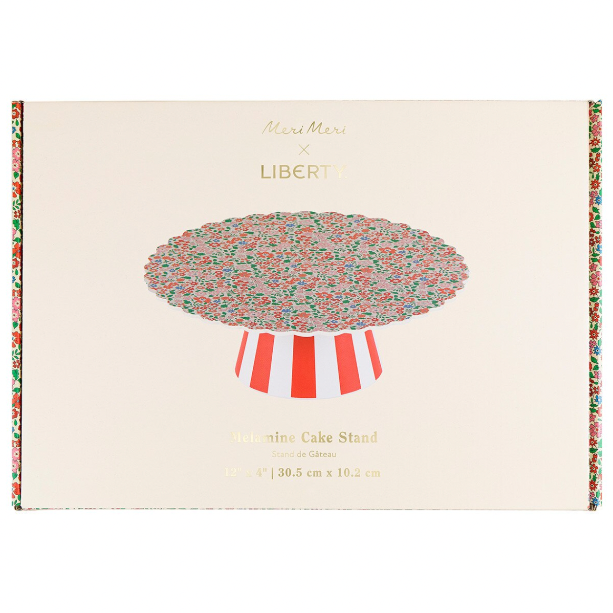 Meri Meri x Liberty Determinine Cake Stand in glass, shown in its packaging box. The box features a cream-colored background with a central illustration of the cake stand holding a colorful floral tablecloth, and decorative borders on the sides.