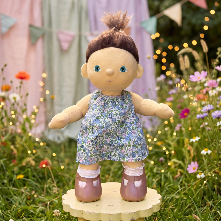 A handmade doll with brown hair, blue eyes, and a blue floral dress, standing on a small wooden platform in a flower garden with string lights.