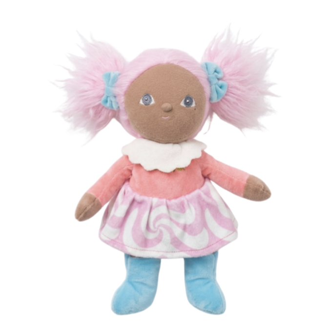 A plush doll with brown skin, blue eyes, and pink hair styled in pigtails with light blue bows. The doll wears a pink dress with a white ruffled collar and a pink-and-white swirl pattern on the skirt, paired with light blue pants, set against a plain white background.