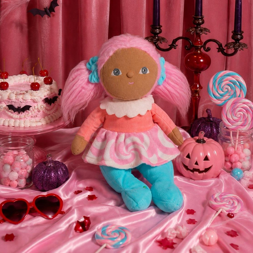 A plush doll with pink hair, a pink and white swirl dress, and blue legs is posed in a Halloween-themed setup with candles, lollipops, a jack-o'-lantern, candy jars, and a pink curtain backdrop.