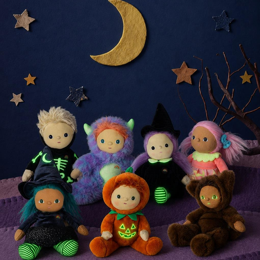 Six plush Halloween-themed dolls arranged on purple steps against a dark blue starry night backdrop with a gold crescent moon. The dolls wear costumes in black, purple, orange, brown, pink, and green.