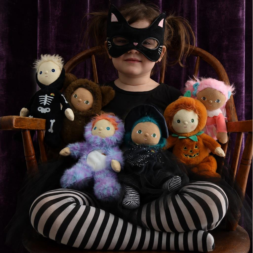 A child wearing a black cat-eye mask and black-and-white striped tights sits cross-legged on a wooden chair, holding several small plush dolls against a dark purple velvet curtain background.