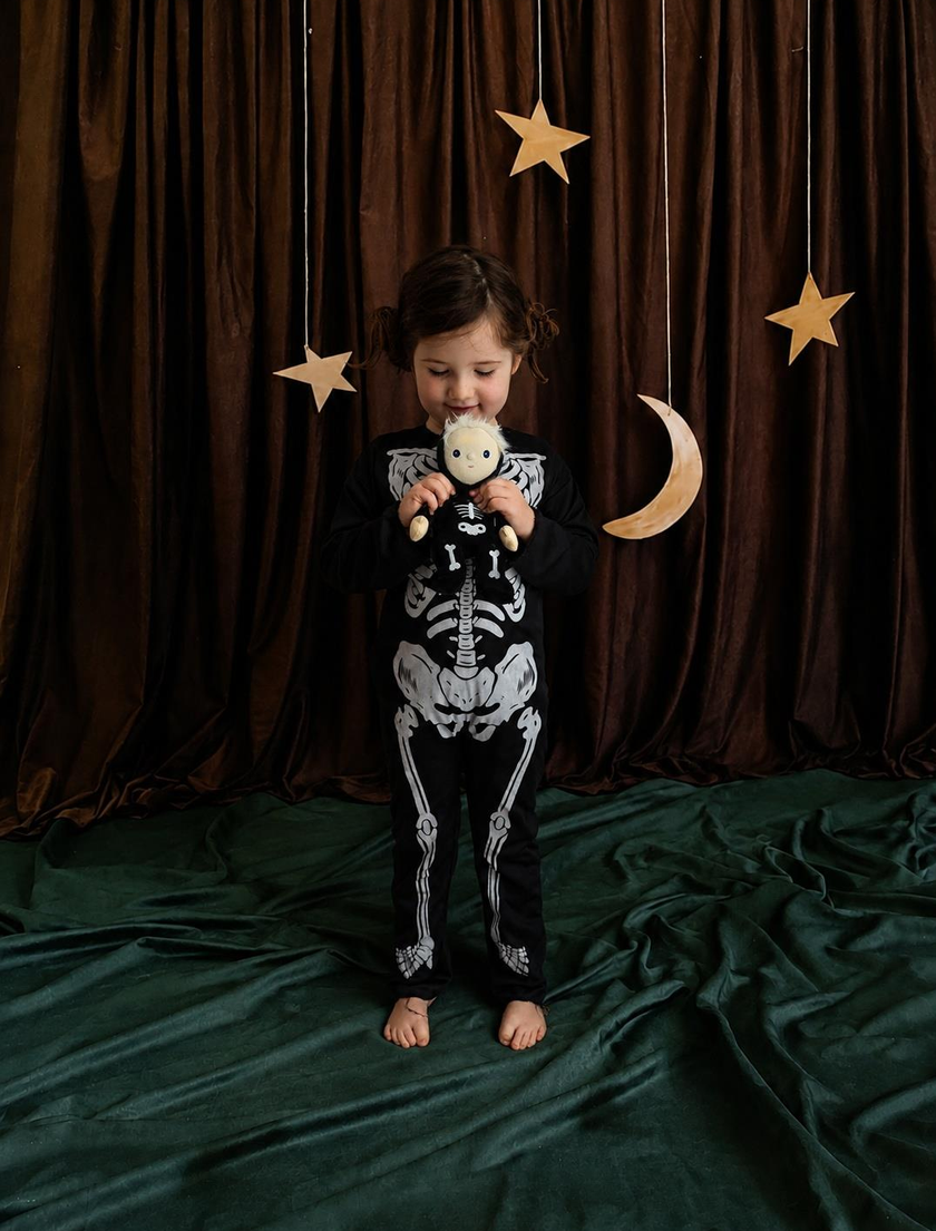 A young child stands barefoot in front of a dark brown curtain backdrop, wearing a black long-sleeve pajama set with a white skeleton print. The child is holding a small plush toy and is posed against a decorated background with hanging wooden stars and a crescent moon.