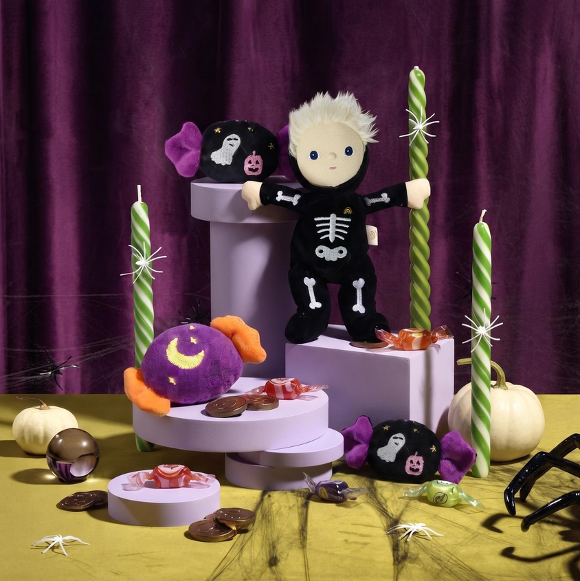 A Halloween-themed styled product display featuring a plush doll dressed in a black skeleton outfit, surrounded by candy corns, a purple moon-themed plush, black ghost plushies, green-and-white striped candles, and spider decorations on a yellow surface against a dark purple curtain backdrop.