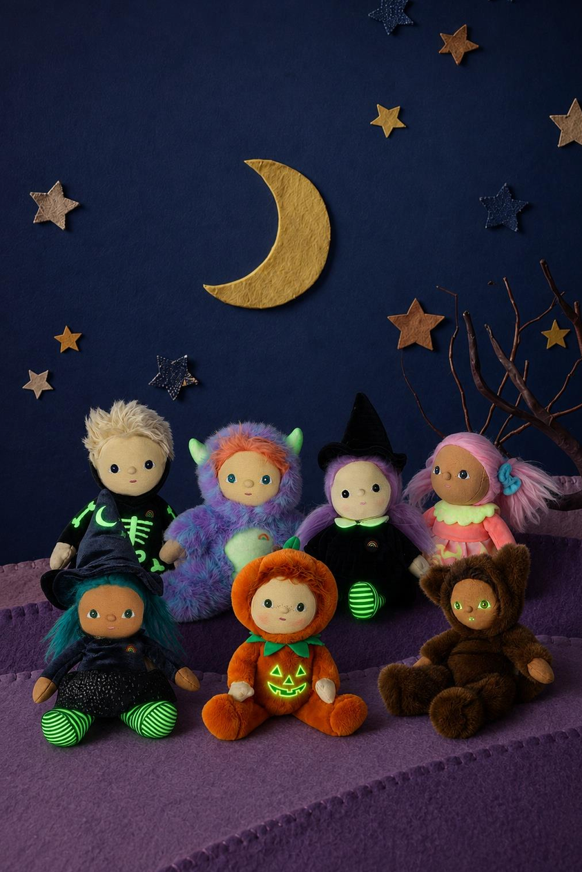 Six plush Halloween-themed dolls arranged on purple steps against a dark blue starry night backdrop with a gold crescent moon. The dolls wear costumes in black, purple, orange, brown, pink, and green.