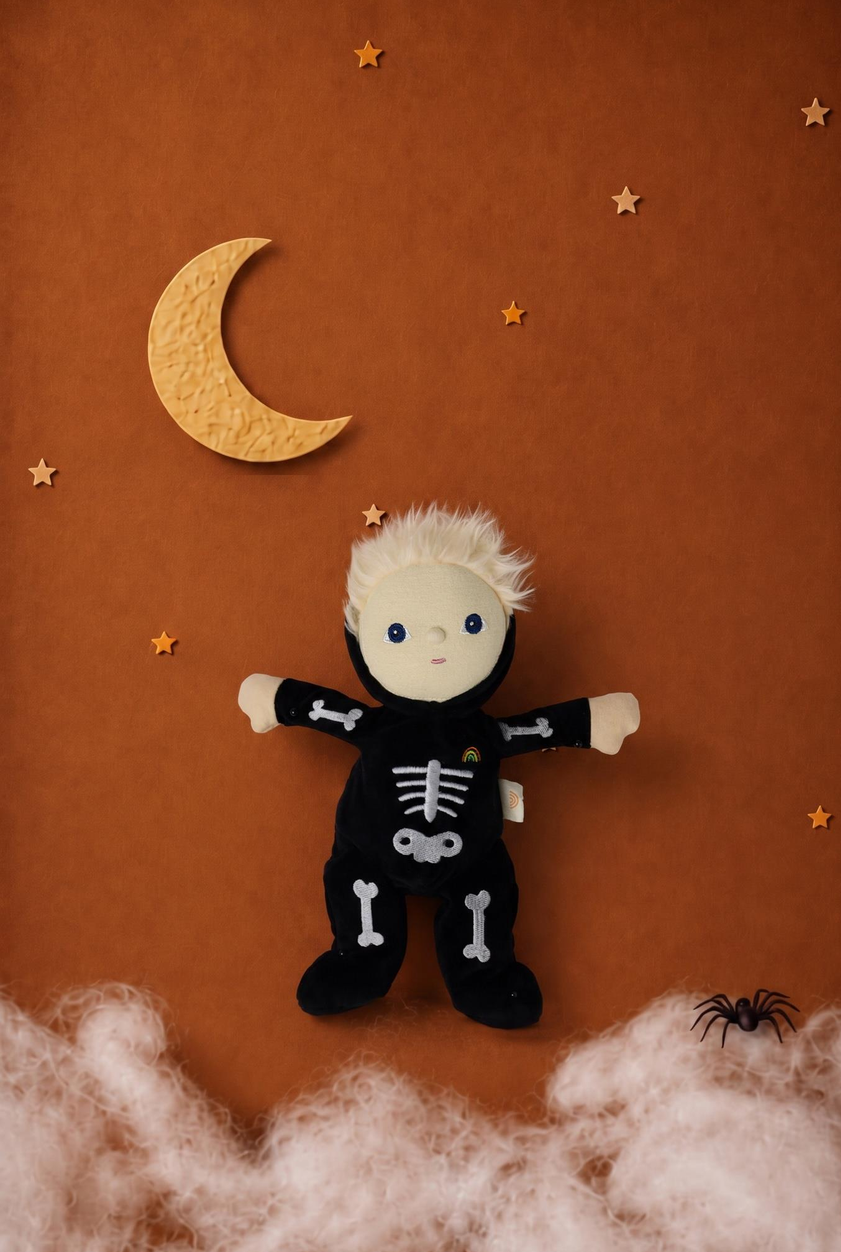 A plush toy doll dressed in a black skeleton costume with white bone details, posed against a warm brown background with a yellow crescent moon, stars, cotton clouds, and a small black spider decoration.