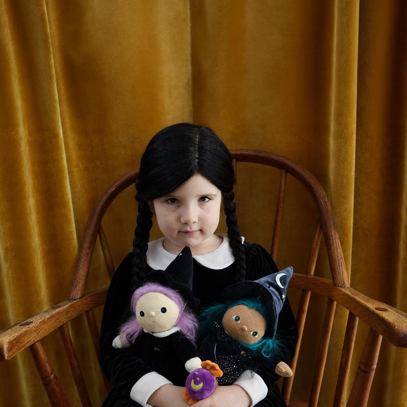 A young girl with black braids sits in a wooden chair against a gold velvet curtain, wearing a black dress with a white collar and holding two small witch-themed dolls.