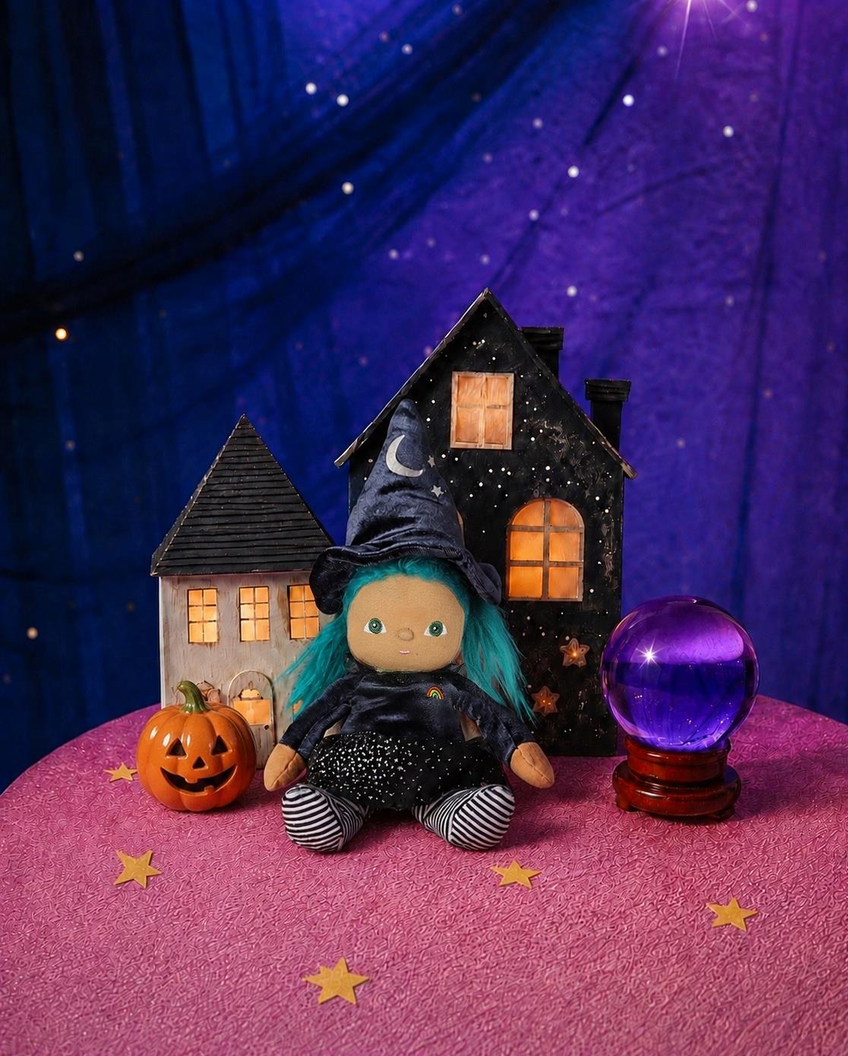 A Halloween-themed doll with blue hair sits between two small toy houses, with a glowing purple crystal ball and a jack-o'-lantern on a pink surface against a purple starry backdrop.