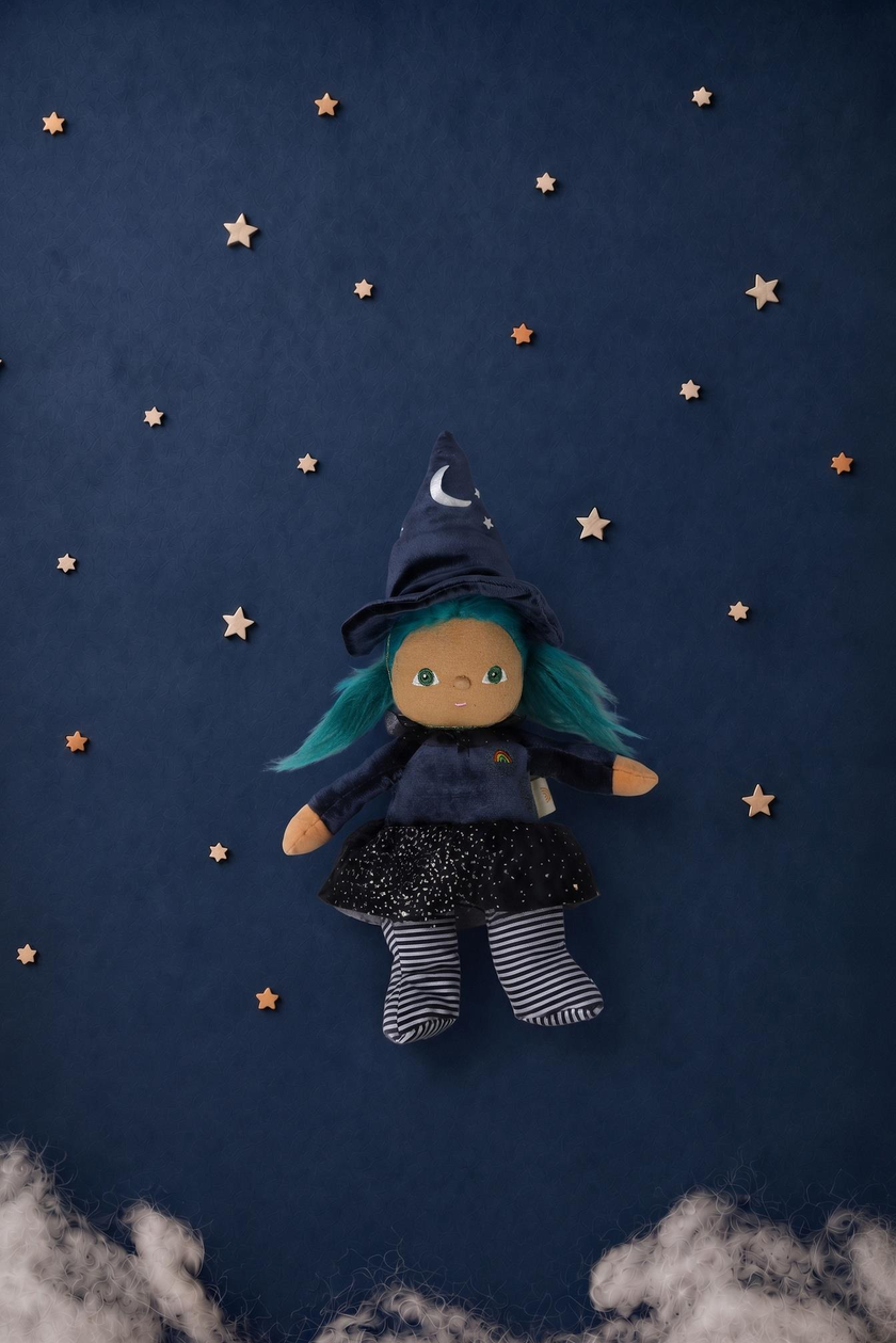 A witch doll with teal hair, a navy blue hat with a crescent moon, a dark dress, and striped tights, floating against a dark blue starry background with small wooden stars and white cloud accents.