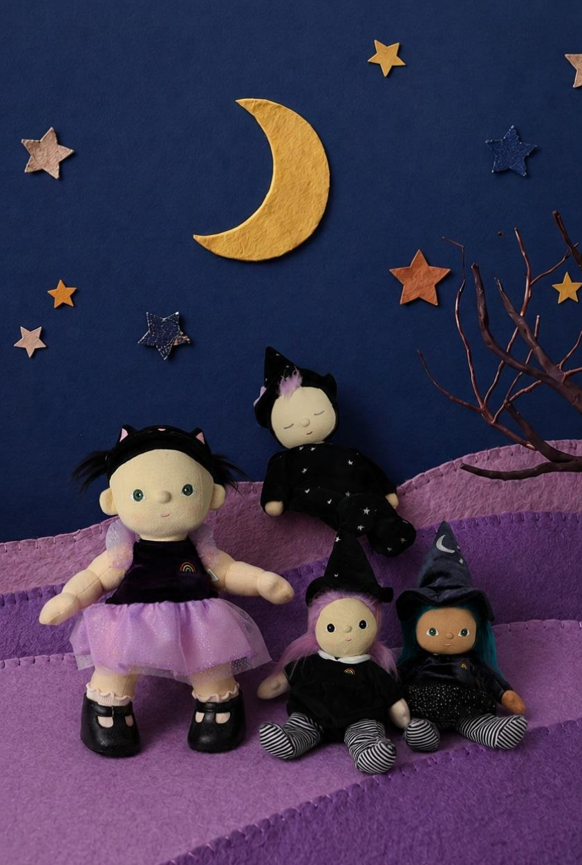 Four witch-themed plush dolls arranged on purple steps against a dark blue fabric sky backdrop with a gold crescent moon, stars, and bare branches.