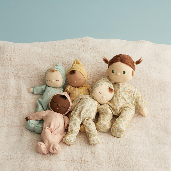 Five handmade fabric baby dolls with diverse skin tones and hair colors, dressed in pastel and floral one-piece outfits, arranged on a soft beige textured blanket against a light blue background.
