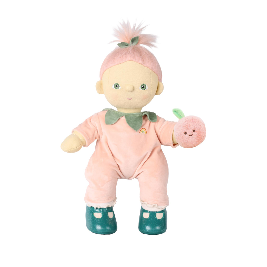 A plush doll with pale skin, green eyes, and pink hair styled in a small topknot. The doll wears a light pink one-piece outfit with a green collar and holds a round pink plush apple.