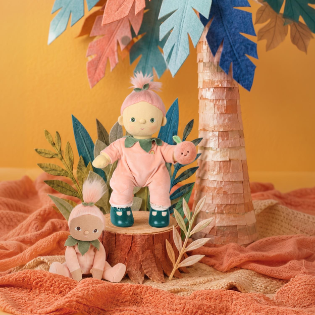 A styled product display featuring two soft fabric dolls in pastel pink outfits, one standing on a small wooden stump and one seated on a textured peach blanket. The scene is decorated with a paper palm tree, colorful leaves, and green foliage against a warm yellow-orange background.