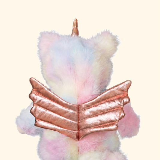 A plush unicorn toy viewed from the back, featuring pastel pink, blue, and white fur, a metallic rose gold horn, and metallic rose gold wing-like back details, set against a solid pale cream background.