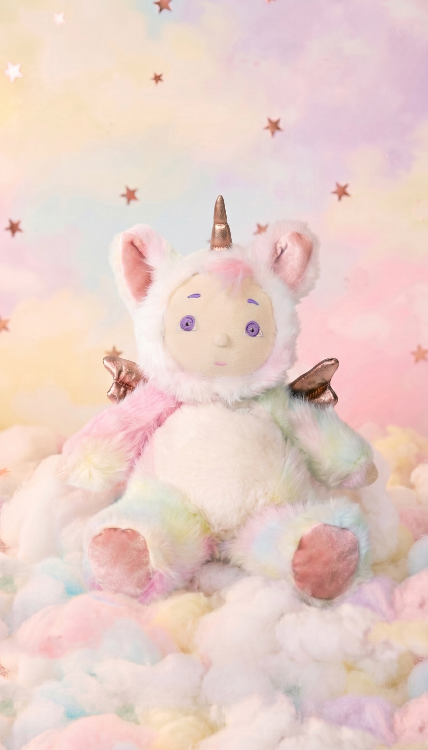 A plush unicorn toy with pastel rainbow fur, pink inner ears, a metallic gold horn, and small metallic wings, sitting on soft pastel clouds against a dreamy pastel sky background with star accents.