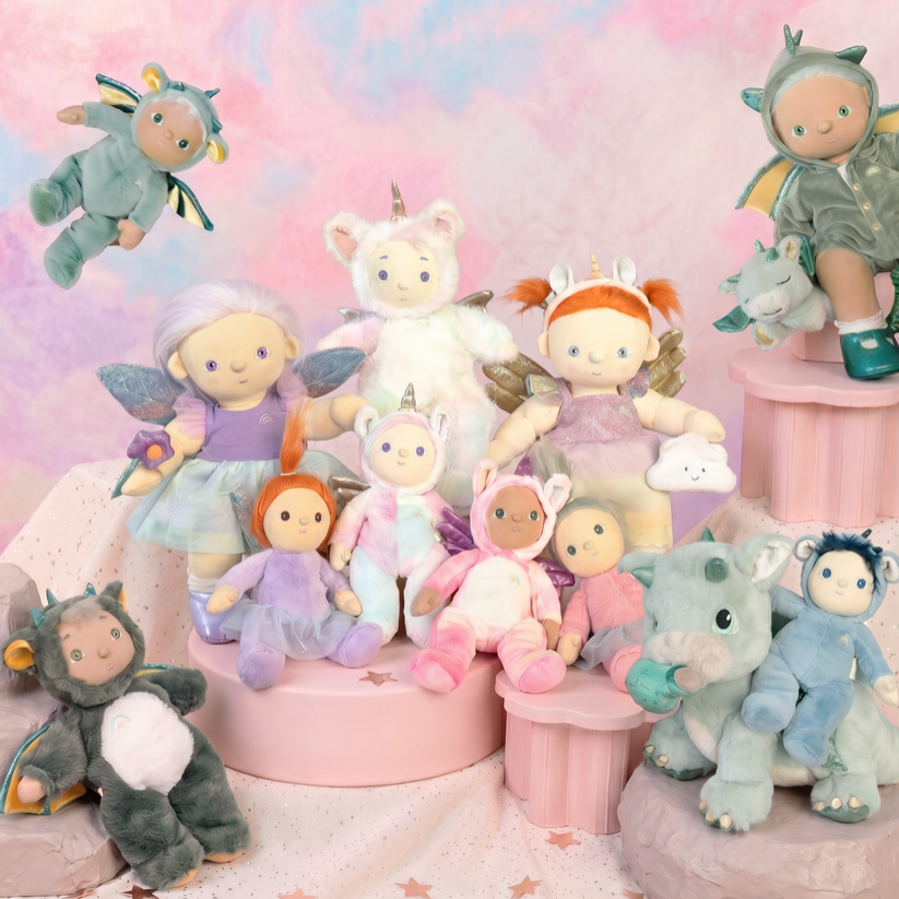 A group of pastel-colored plush toys and dolls posed together on pink display pedestals against a soft pink and blue gradient backdrop. The toys include unicorns, fairies, dragons, and small animal characters in coordinated pastel outfits.