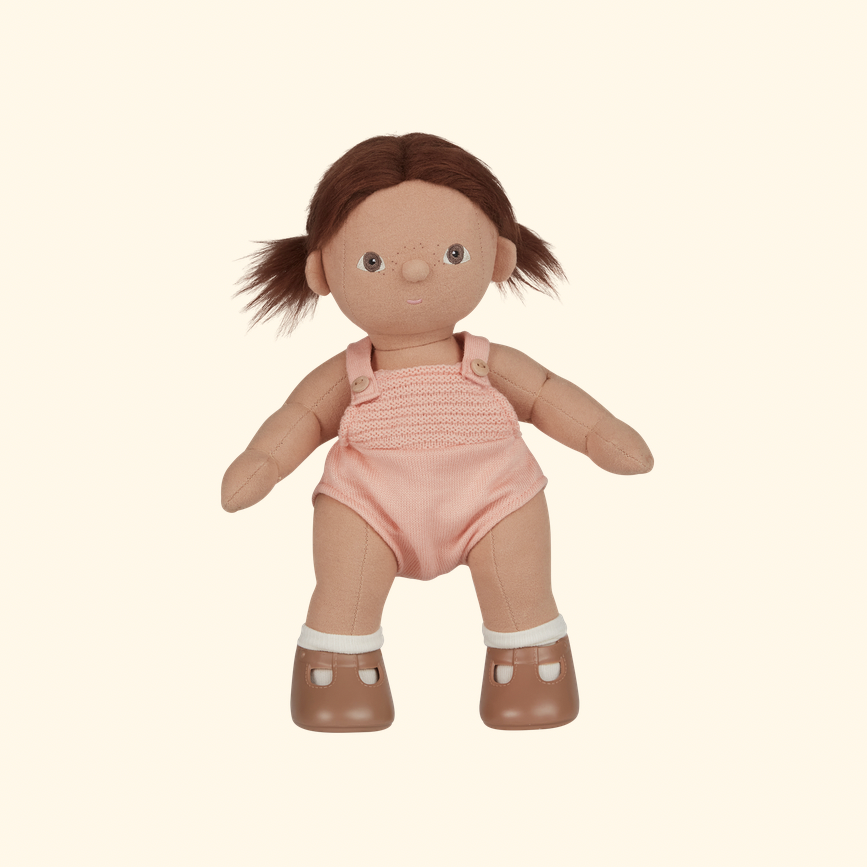 A soft plastic doll with brown hair in pigtails, wearing a pink knit romper and brown shoes with white socks, shown against a plain cream background.