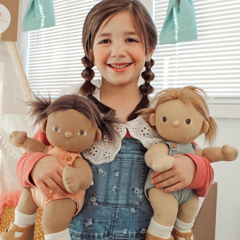 A smiling young girl with braided hair holds two plush dolls dressed in colorful clothing, posed in front of window blinds with soft natural light.
