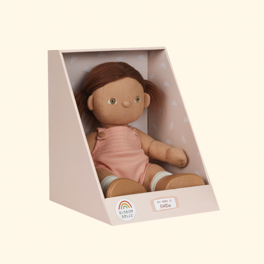 A soft fabric doll with brown hair and a pink outfit displayed inside a pale pink triangular presentation box. 