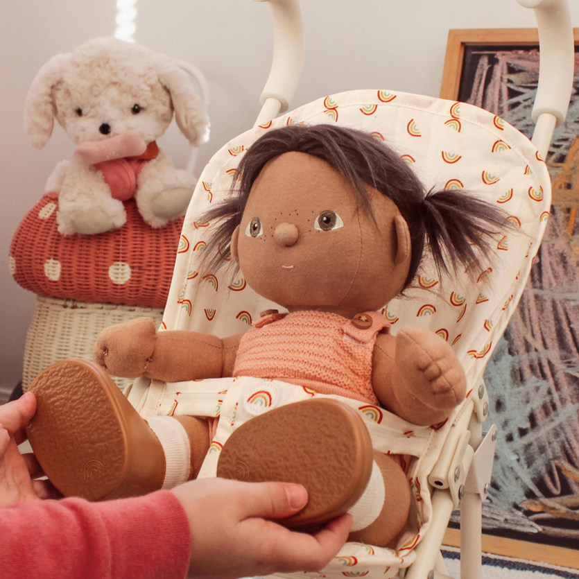 A plush doll with dark hair sits in a cream-colored fabric stroller with a rainbow pattern, held by hands. A plush sheep toy is visible in the background on a wicker basket.