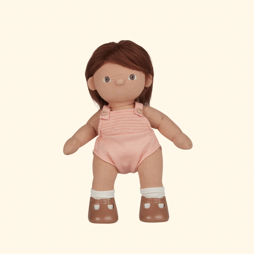 A doll with brown hair, freckles, and a pink knit romper, standing against a solid cream background.