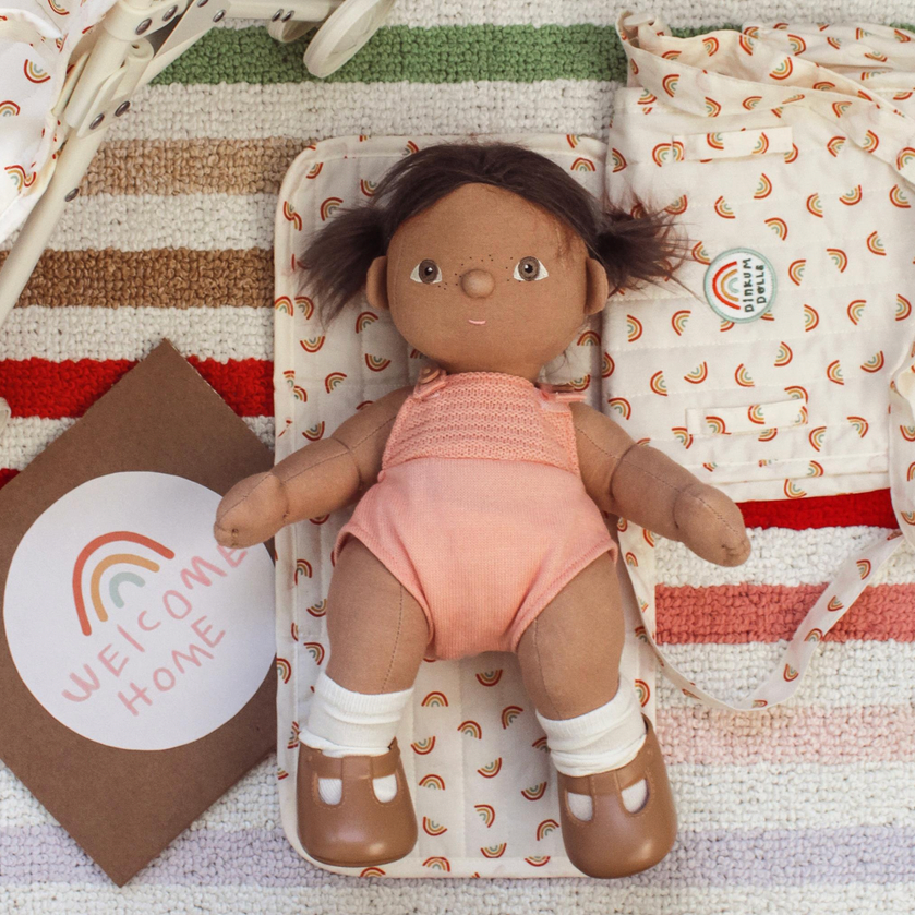 A doll with dark brown hair in pigtails lies on a striped blanket with a peach bodysuit, white socks, and brown shoes. A matching patterned pouch and a brown card reading 'WELCOME HOME' with a rainbow are arranged around it.