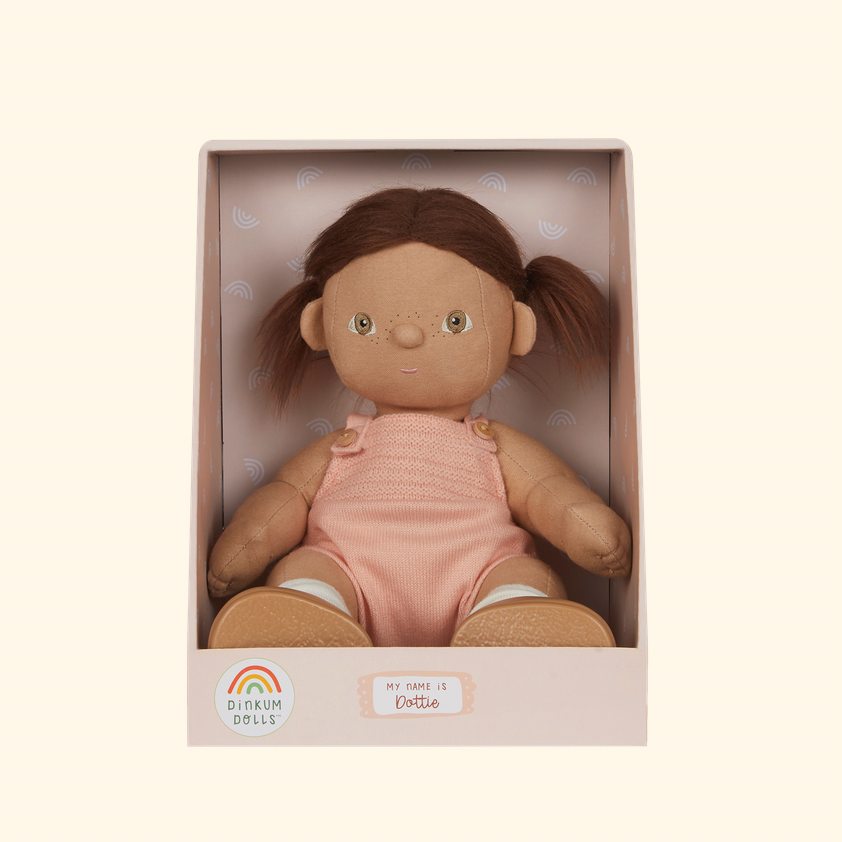 A boxed plush doll with brown hair in pigtails, wearing a pink romper, displayed inside a pale pink box 
