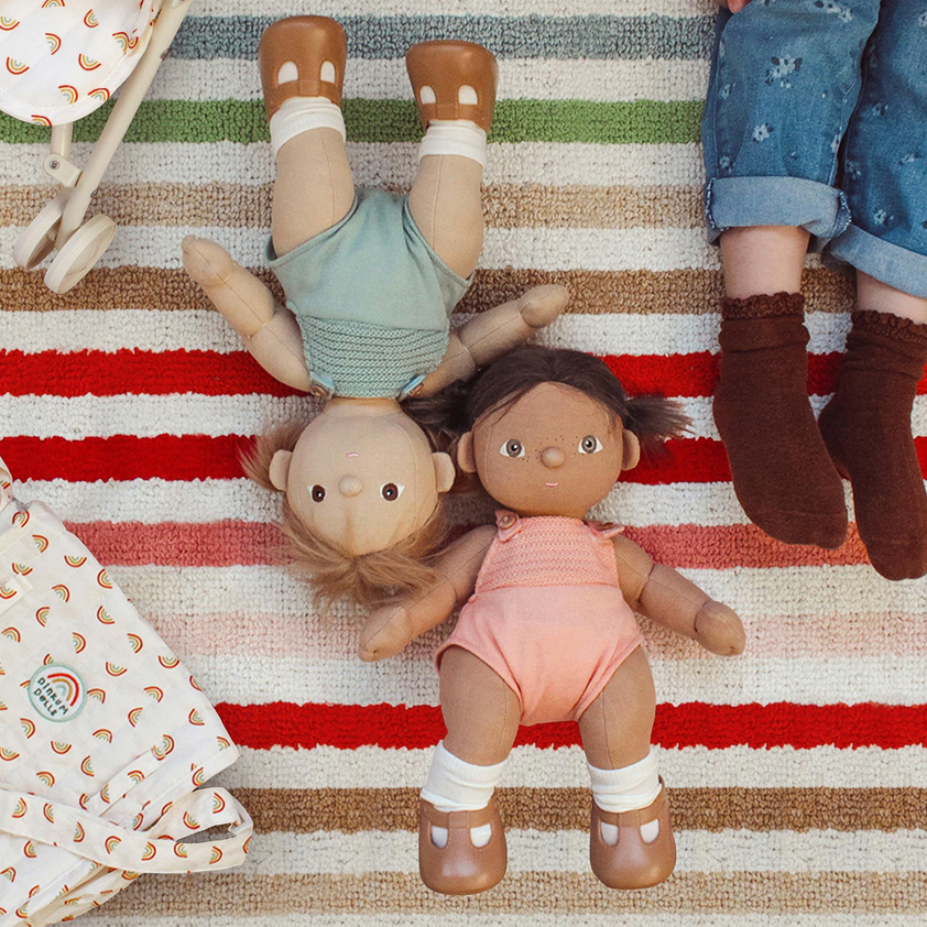 Two vintage-style dolls are arranged on a striped knit blanket with other toys partially visible around them. One doll is upside down wearing a light blue outfit, and the other stands facing up in a peach bodysuit with brown shoes.