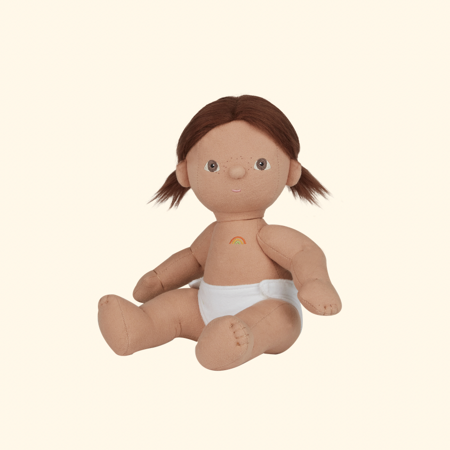 A soft fabric baby doll with brown hair in pigtails, a tan body, and a white diaper, seated against a plain warm cream background.