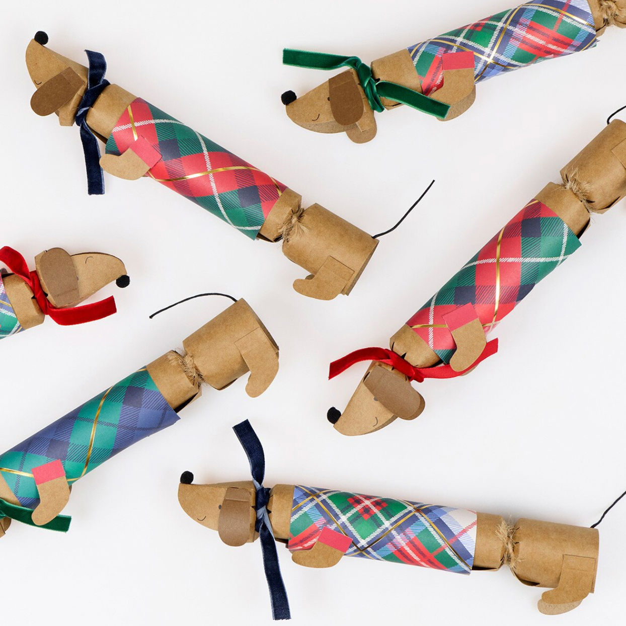Five dachshund-shaped cracker ornaments made from brown cardboard tubes, wrapped in colorful tartan plaid paper, and decorated with ribbon collars, arranged on a plain white background.