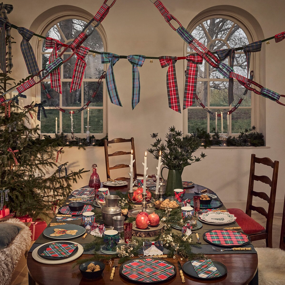 A styled Christmas dining room scene with tartan bunting garlands strung across arched windows, a decorated tree, candles, and matching plaid tableware and napkins on a wooden table.