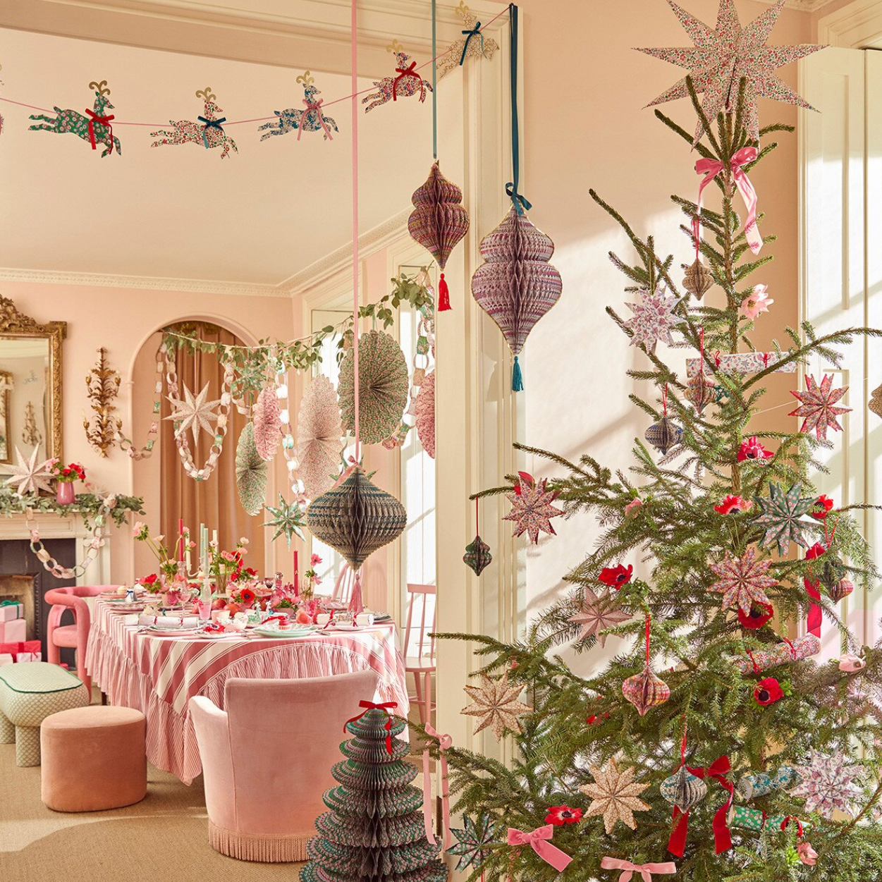 A beautifully decorated Christmas tree in a warm, pink-toned dining room. The tree is adorned with pink and silver ornaments, ribbons, and hanging paper lanterns, alongside a pink tablecloth, blush chairs, and festive garlands.