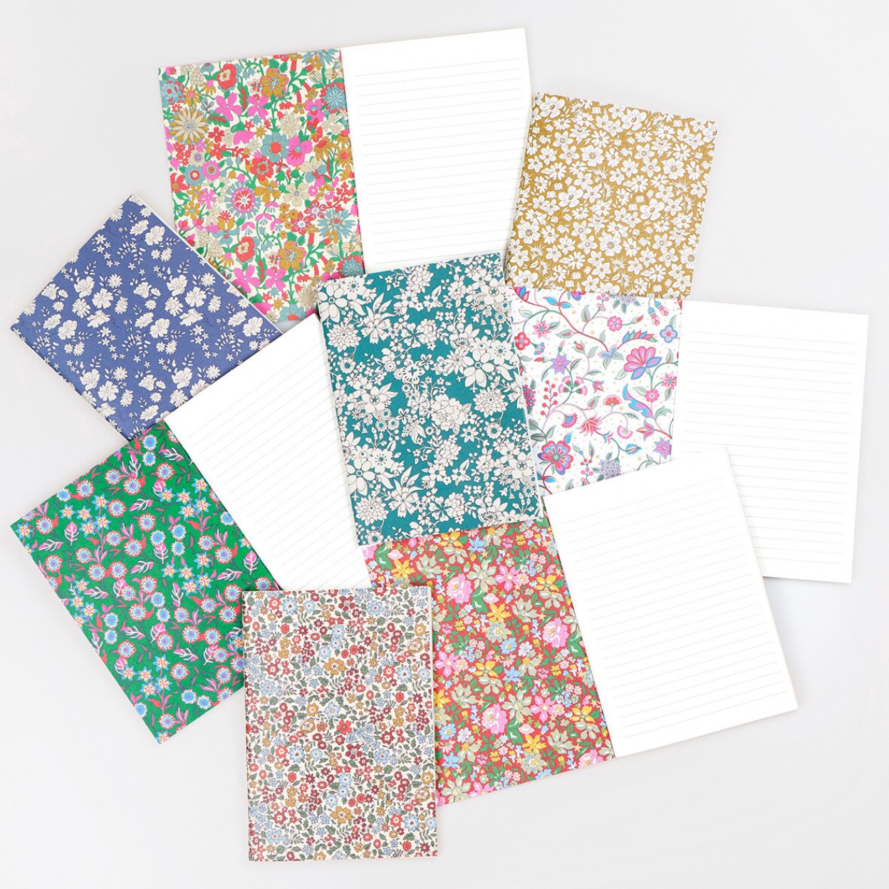 A flat lay of assorted patterned notepads and lined paper sheets on a white background. The set includes several floral and botanical designs in blue, green, pink, purple, and gold, alongside plain white lined pages.