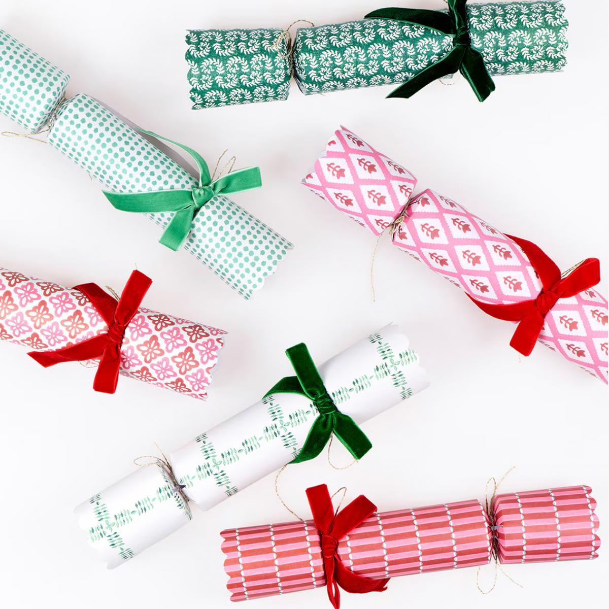 Six Christmas cracker ornaments wrapped in festive patterned paper and tied with red, green, and white ribbons, arranged on a clean white background.